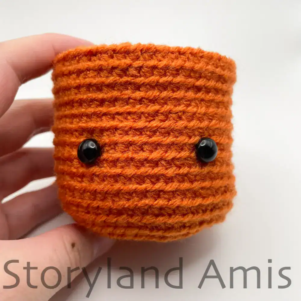 A hand holds up a partially finished crocheted pumpkin, showing where to position the safety eyes.