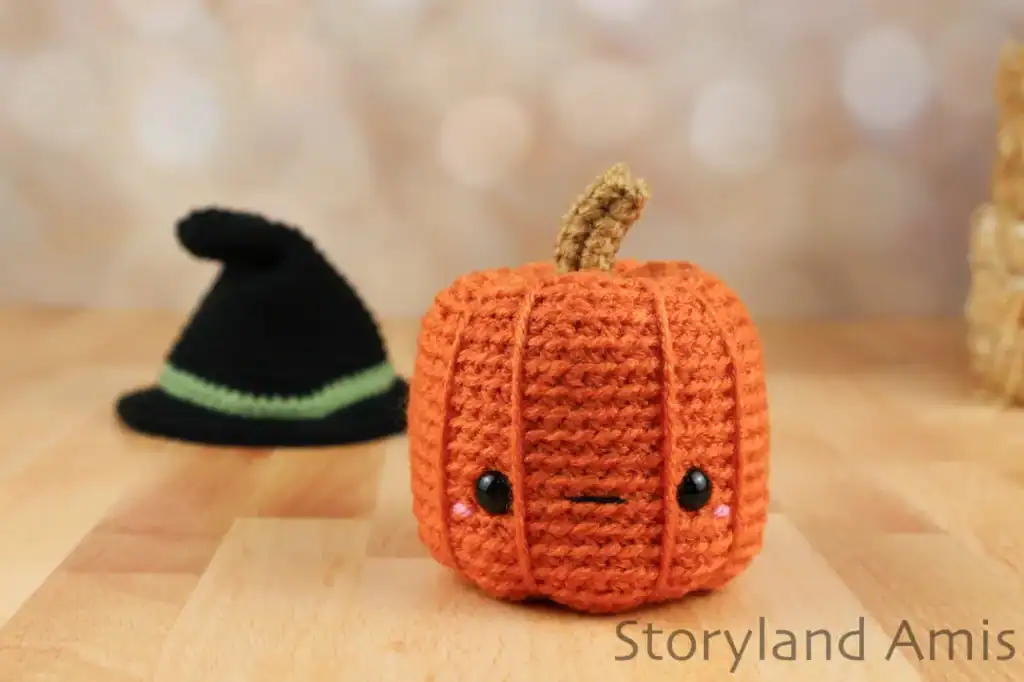 A crocheted pumpkin with a cute face sits on a wooden table. A black witch's hat sits behind it.