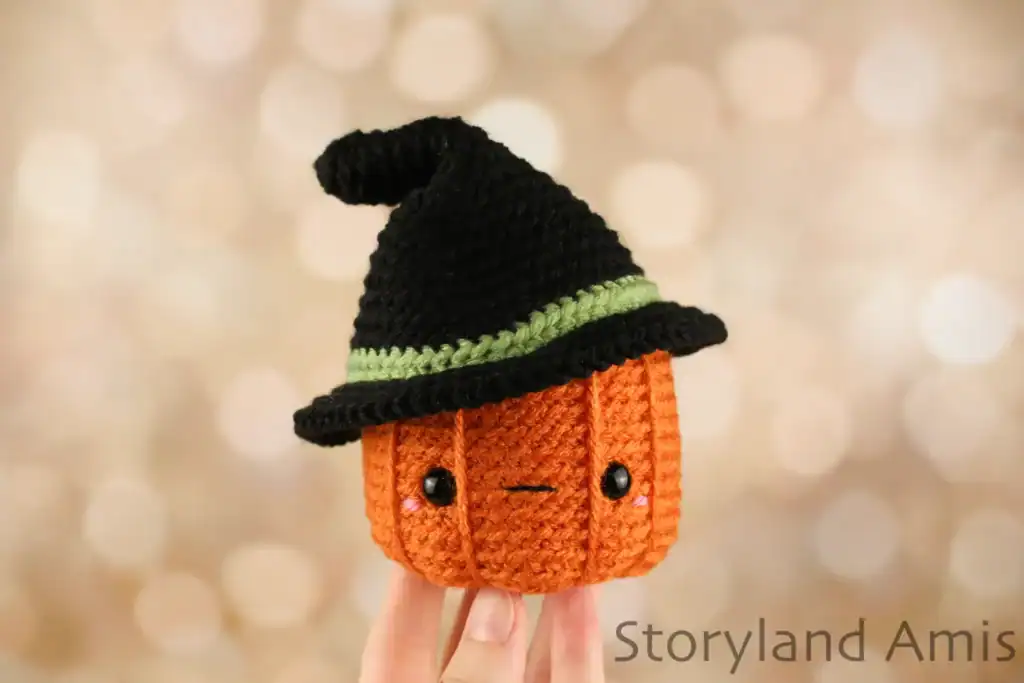 A hand holds up an amigurumi pumpkin wearing a black witch's hat.