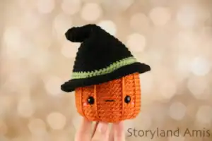 A hand holds up an amigurumi pumpkin wearing a black witch's hat.