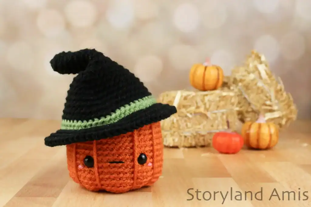 An amigurumi pumpkin with a witch's hat sitting on a wooden table. In the background are mini hay bales and pumpkins.