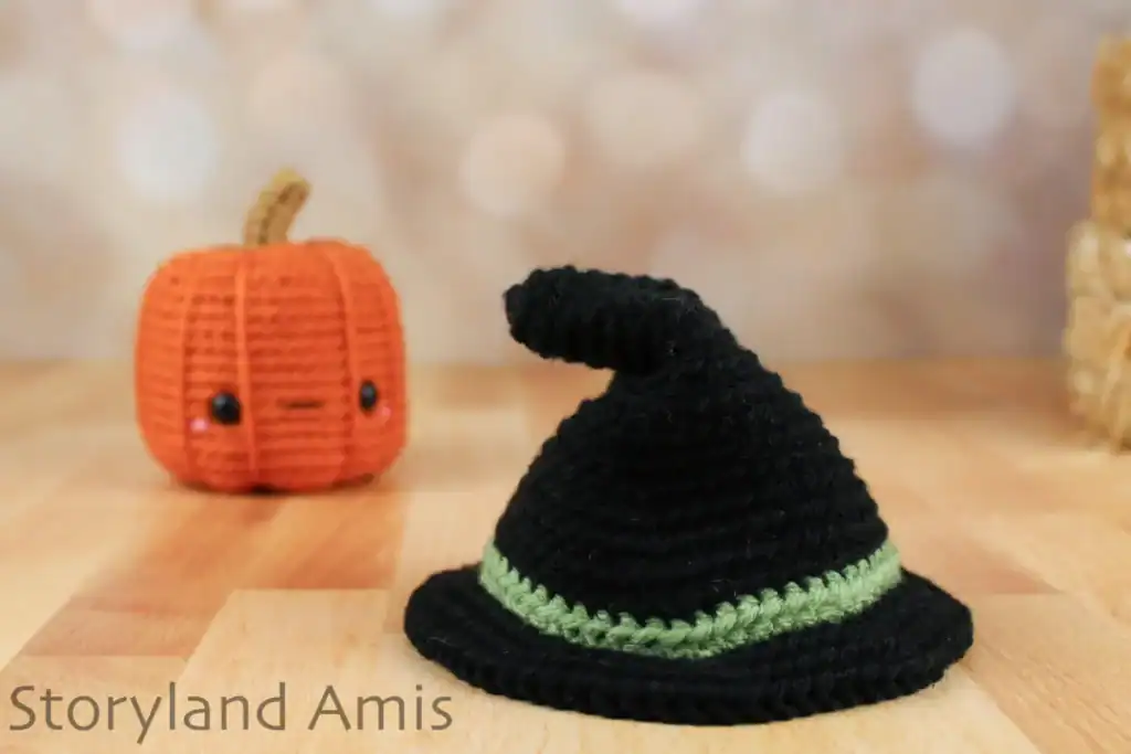 A black witch's hat sits on a wooden table. An amigurumi pumpkin sits behind it.