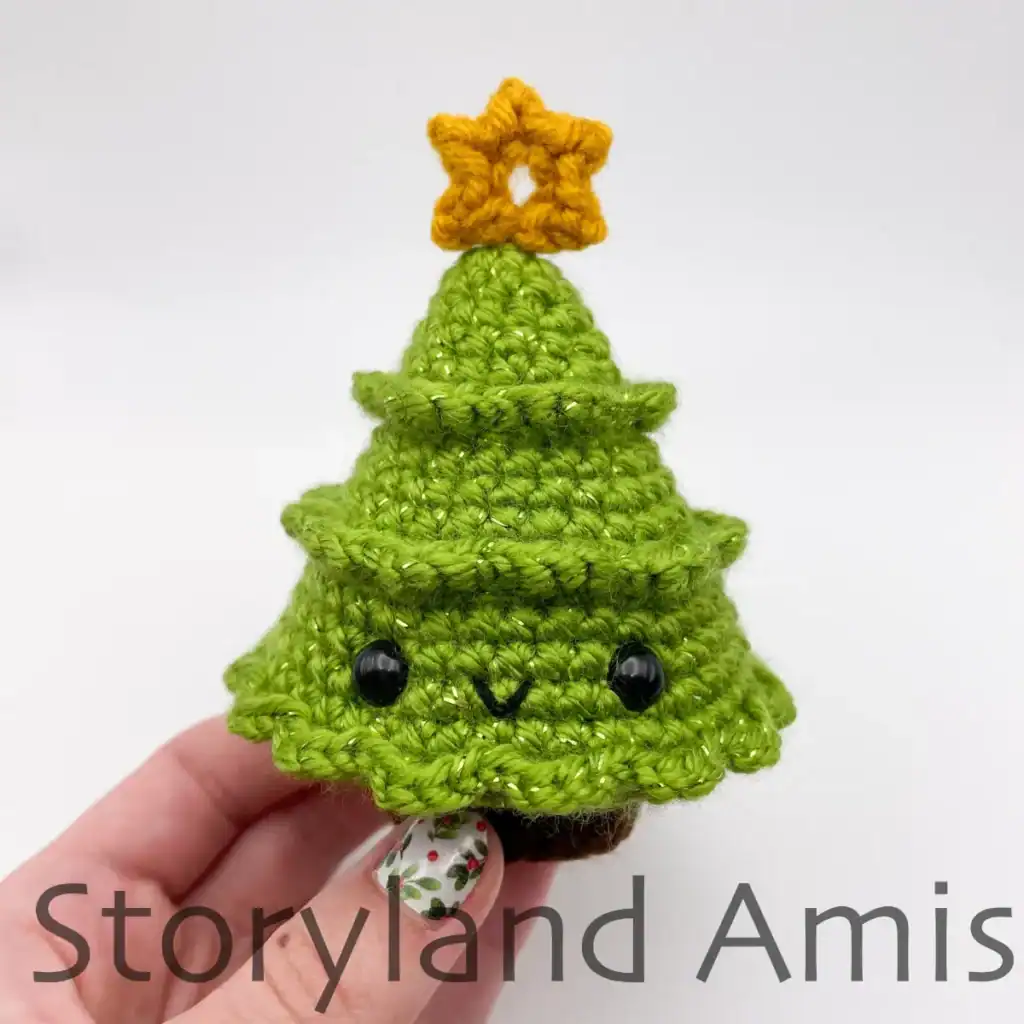 A hand holds up a mini Amigurumi Christmas tree. The green tree has a yellow star on top and a kawaii face.