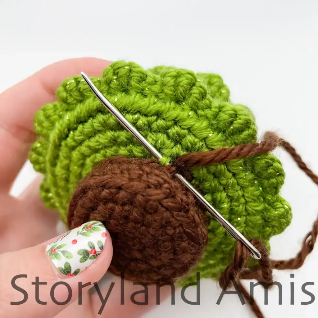 A hand demonstrates how to sew a crocheted trunk to the bottom of an amigurumi Christmas tree.