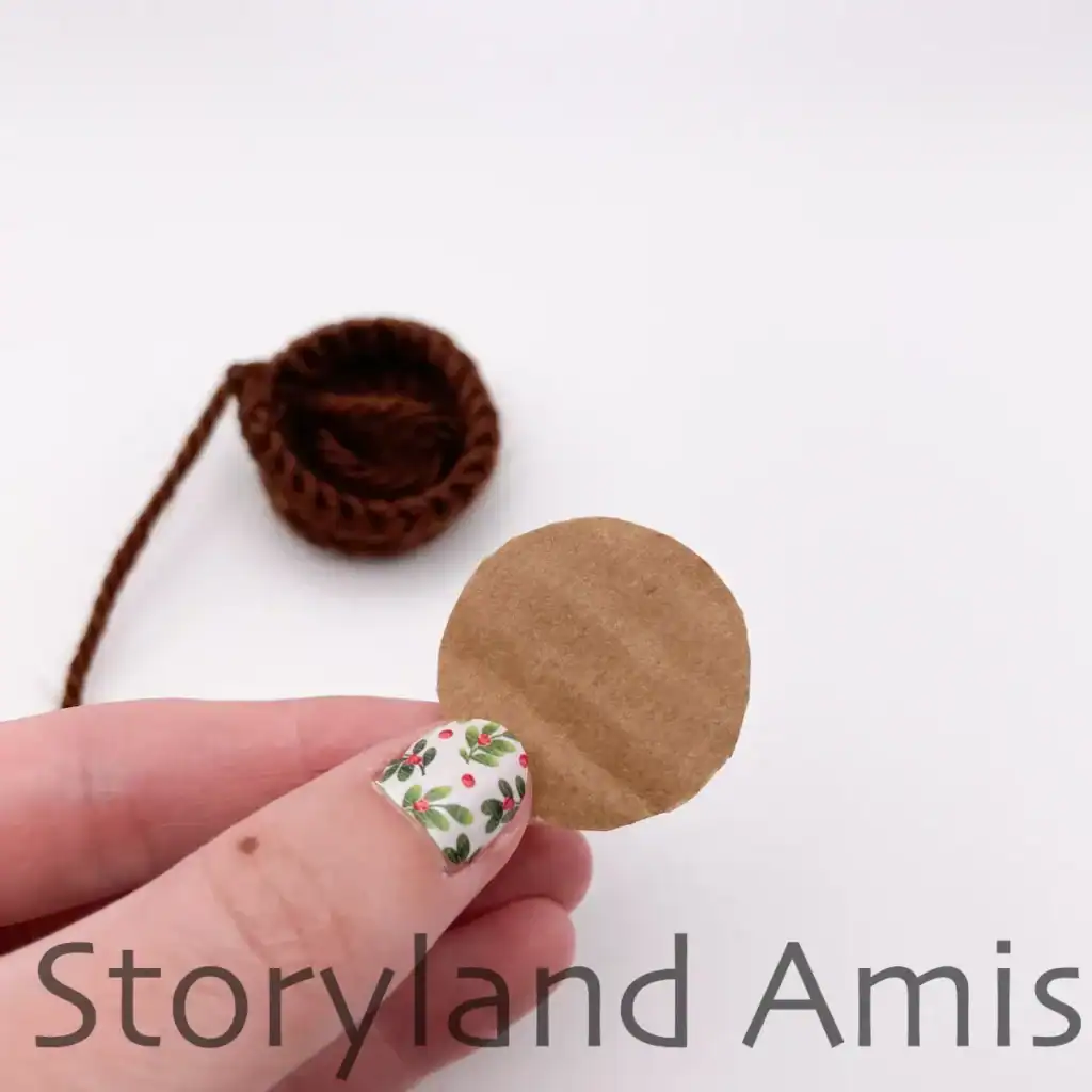 A hand holds a small cardboard circle. In the background sits a crocheted trunk for an amigurumi Christmas tree.