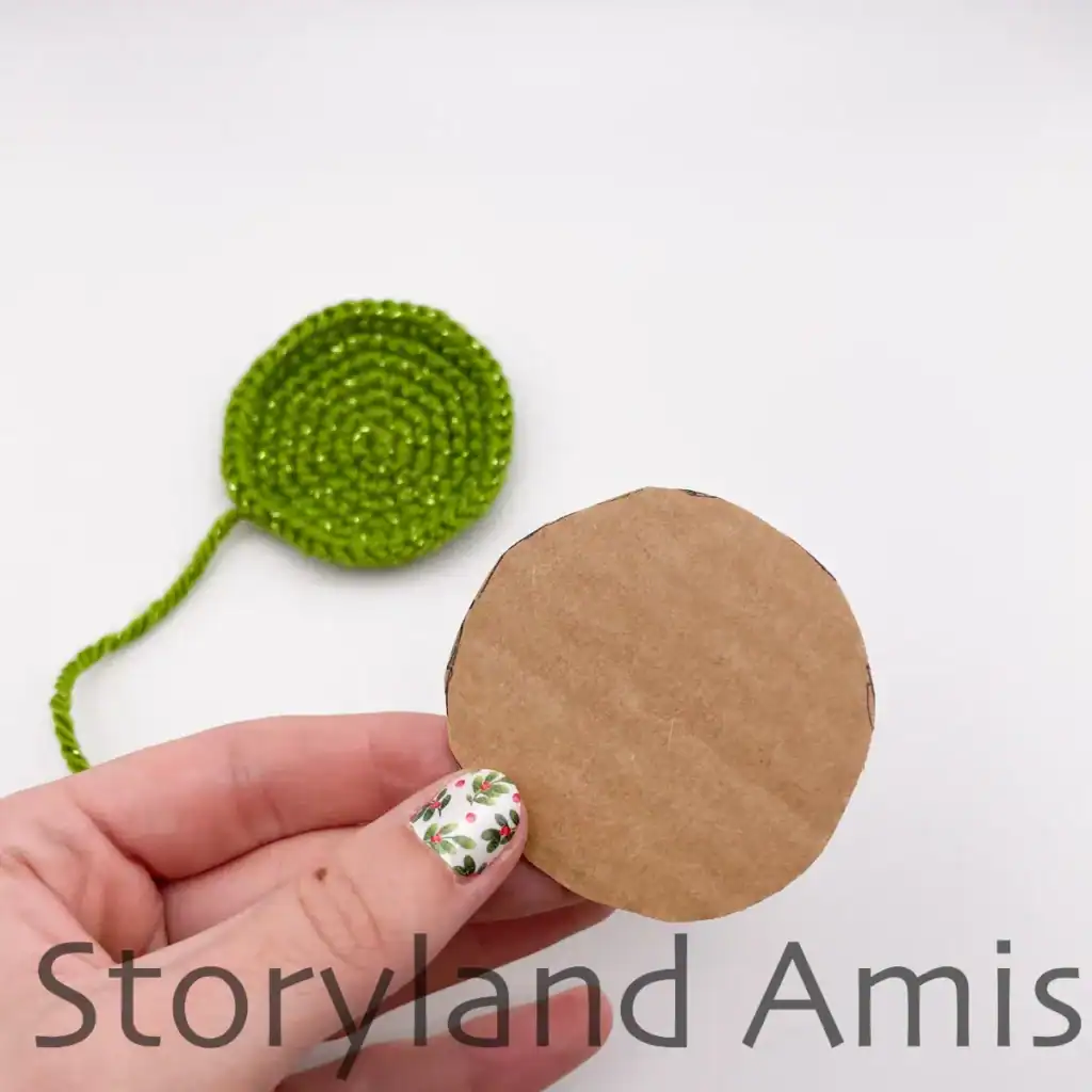 A hand holds up a round piece of cardboard intended to add stability to the bottom of an amigurumi Christmas tree. In the background is the crocheted bottom piece of the Christmas tree.