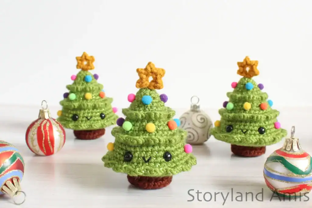Three mini crocheted Christmas trees sit on a wooden table surrounded by colorful Christmas ball ornaments.