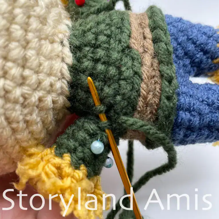 A tutorial photo showing how to sew the arm to the body of an amigurumi scarecrow. A gold-colored tapestry needle is inserted through stitches on the arm and body as it sews them together.
