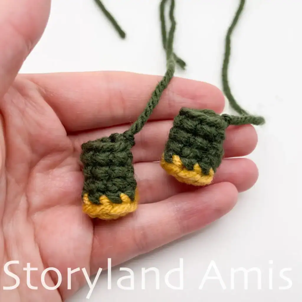 A hand holds two crocheted arms for an amigurumi scarecrow.