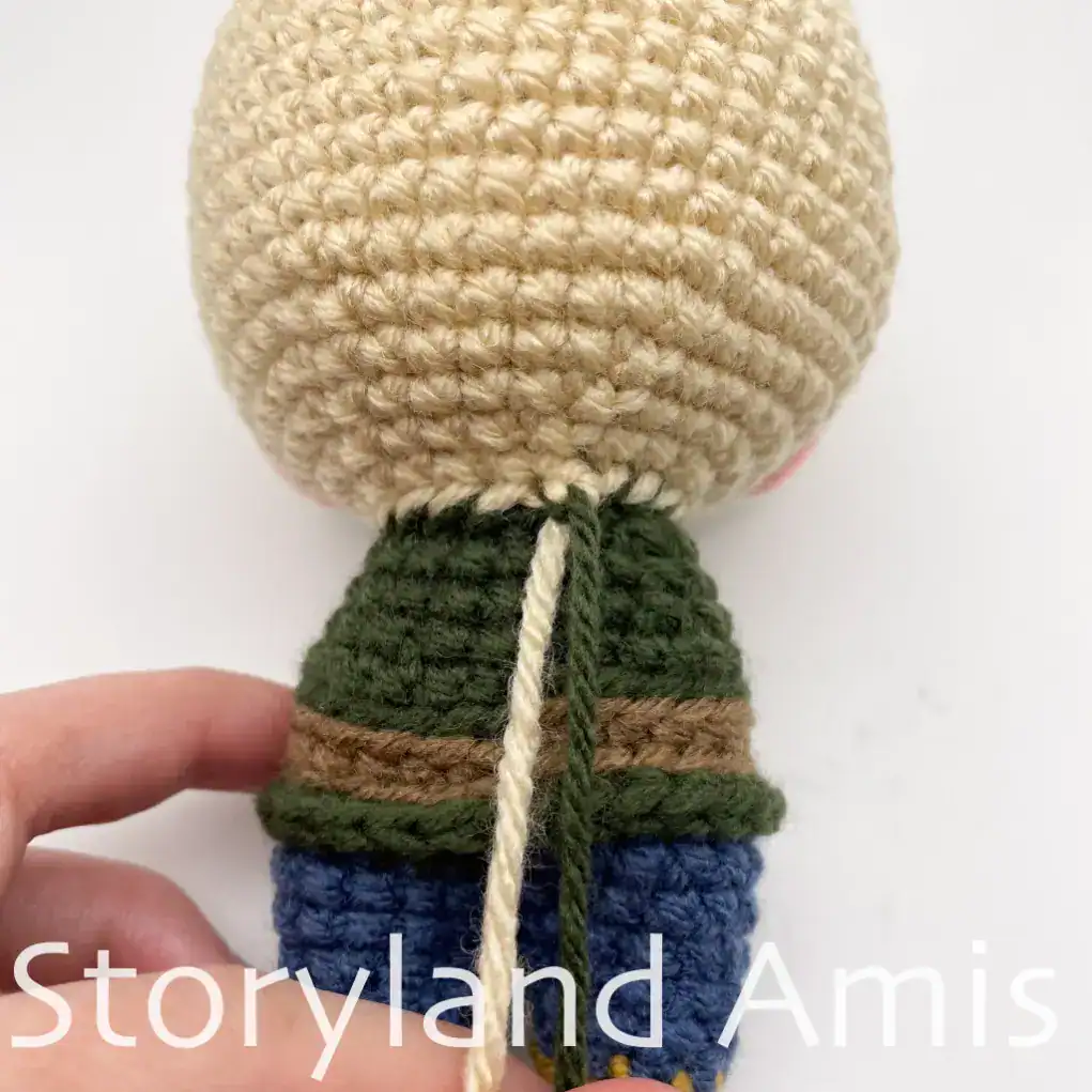A close-up of the back of an amigurumi scarecrow showing the two yarn tails left from the body and head after sewing the two pieces together.