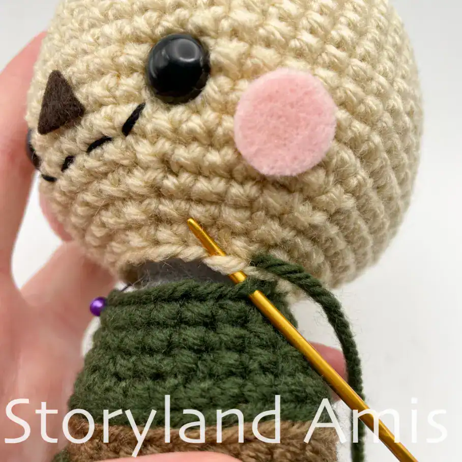 A close-up of an amigurumi scarecrow head being sewn to its body with a gold-colored tapestry needle and yarn tail. A hand holds the doll steady.