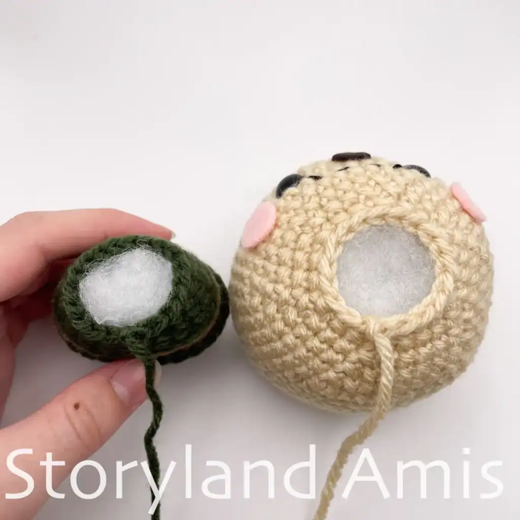 A partially assembled crochet amigurumi scarecrow, showing a beige head with safety eyes, pink felt cheeks, embroidered nose and mouth, and a large open hole at the bottom filled with stuffing. Next to it is a green crochet piece, also partially stuffed, intended to be the scarecrow's body. Both pieces have yarn tails for sewing.