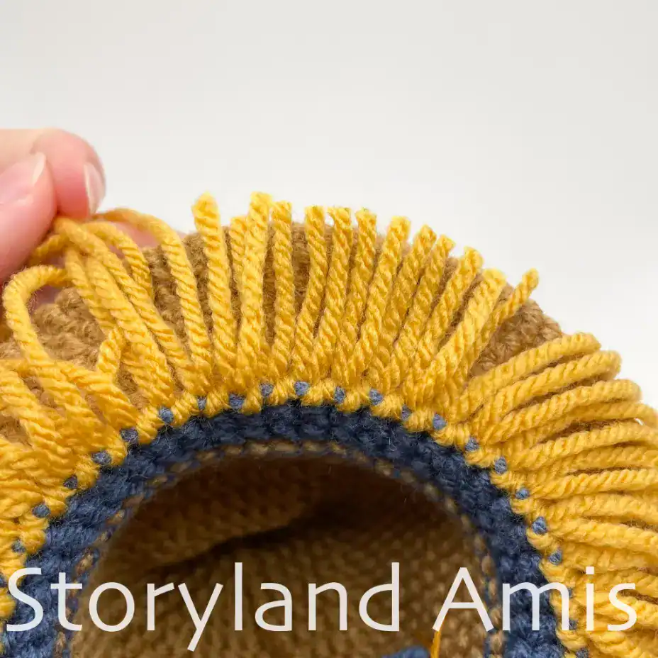 A close-up of the brim of a crocheted scarecrow hat showing the underside of the brim. There is a fringe of yellow yarn around the brim.
