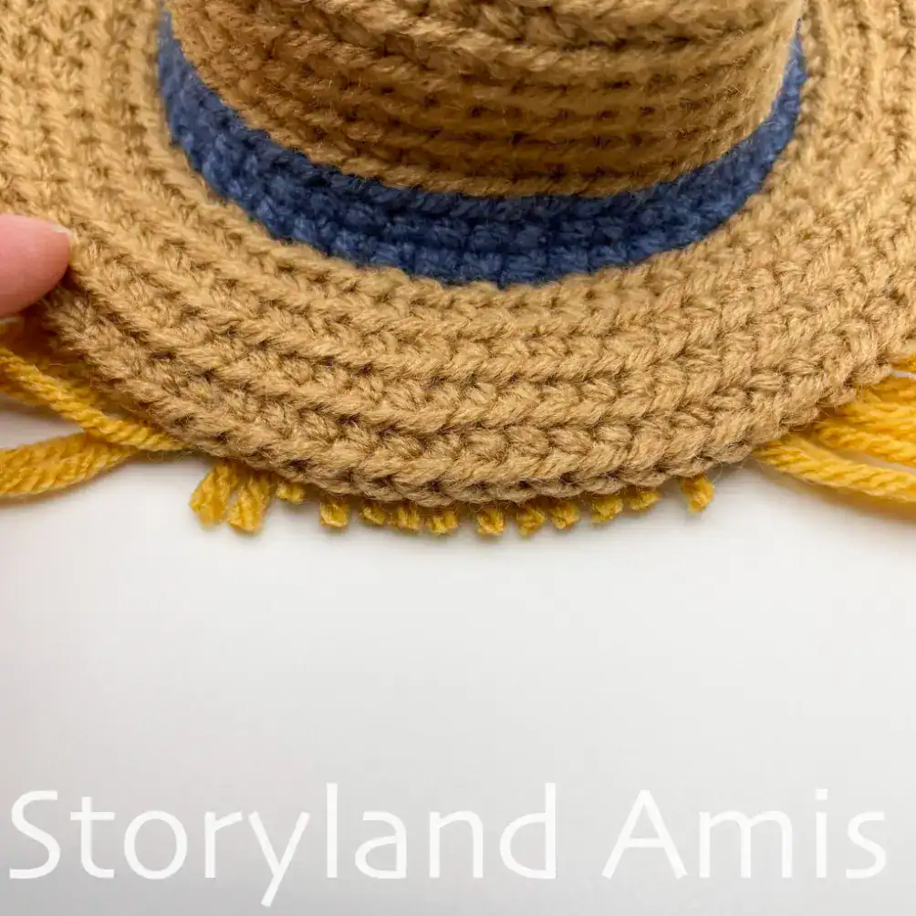 A close-up of the brim of a crocheted hat for an amigurumi scarecrow. The hat is brown with a blue band, and there is a fringe of yarn all around the brim.