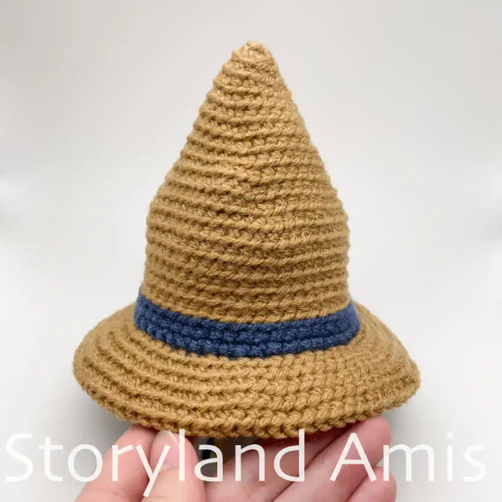 A hand holds a partially finished crocheted hat for an amigurumi scarecrow. The hat is brown with a blue band.