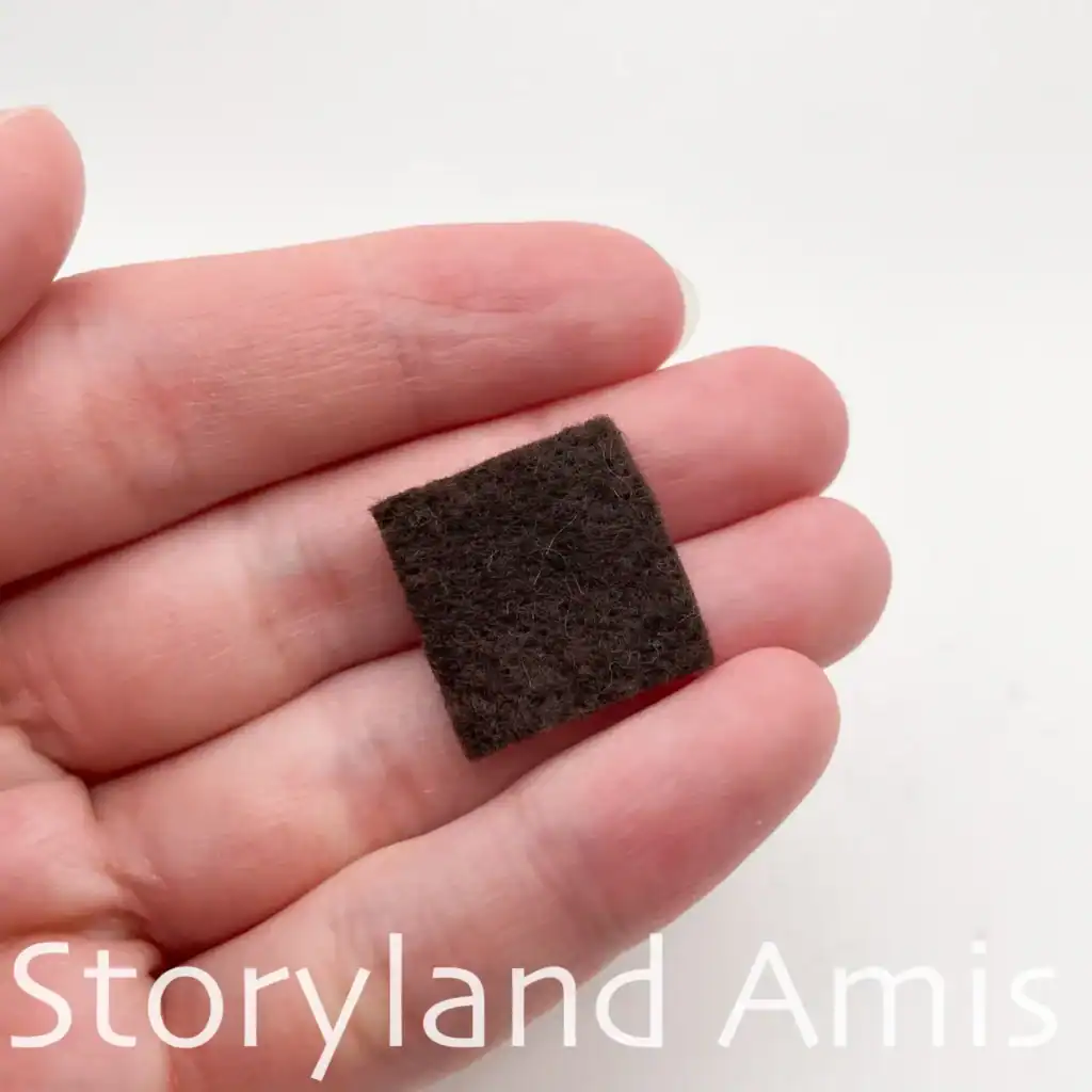 A hand holds a small square of dark brown felt to be used as a patch on a crocheted scarecrow hat.