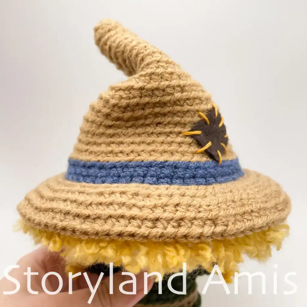 Close-up of a crocheted hat on an amigurumi scarecrow. The hat is brown with a blue band and has a small square brown patch sewn to the side.