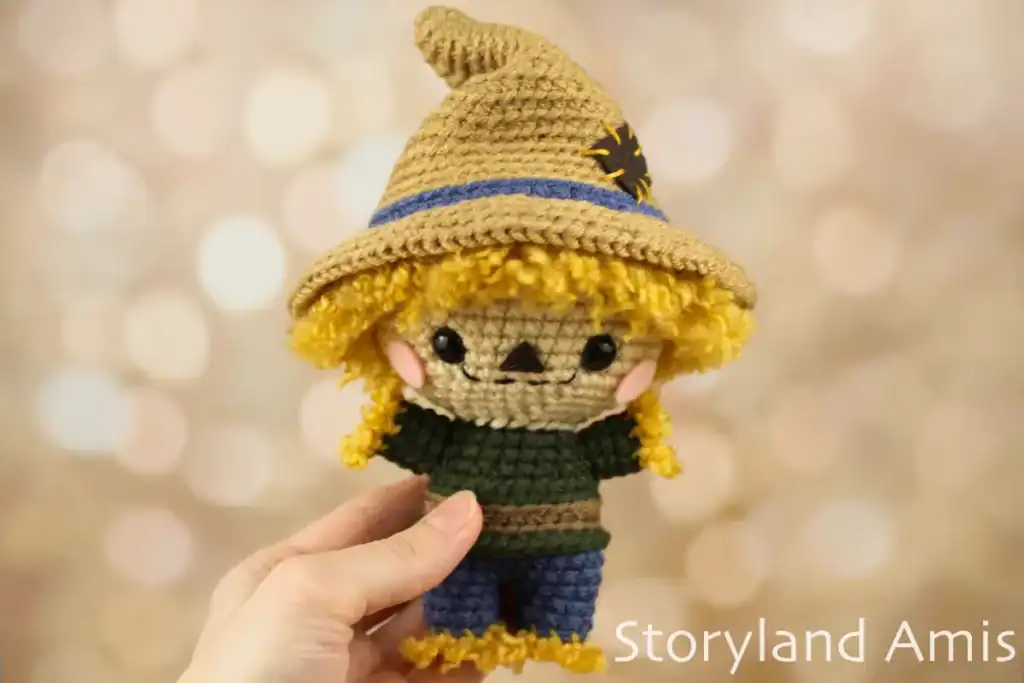 A hand holds a crocheted scarecrow. The scarecrow has a brown colored hat and a green shirt with blue pants.