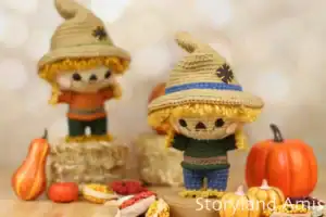 Two handmade amigurumi scarecrows with curly yellow hair and large tan hats stand on small hay bales surrounded by mini pumpkins, colorful dried corn, and fall decorations. One scarecrow wears a green sweater with blue pants, and the other has an orange sweater with green pants. Both have stitched smiles and rosy cheeks. The cozy autumn scene is softly lit with a blurred background.