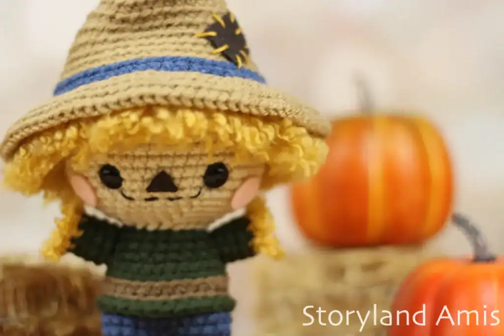A close-up of a crocheted scarecrow. There are small hay bales and pumpkins in the background.
