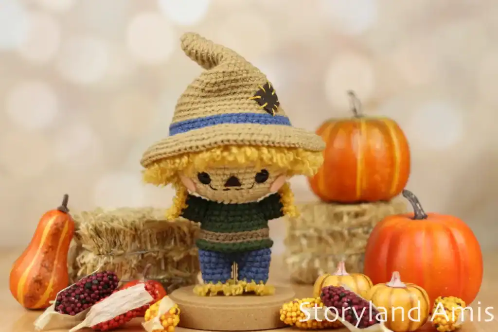 An amigurumi scarecrow with small hay bales, pumpkins, and corn in the background and foreground.