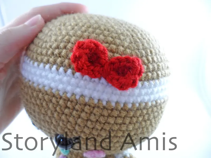 A close-up of the side of an amigurumi gingerbread girl's head showing a red bow sewn to the side of the head.