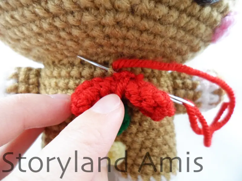 A close-up of the front of an amigurumi gingerbread boy's body. A red crocheted bowtie is being sewn to the front of the body using a silver tapestry needle.