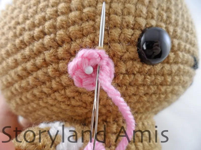 A close-up of a pink crocheted cheek being sewn onto the face of an amigurumi gingerbread boy using a silver tapestry needle.
