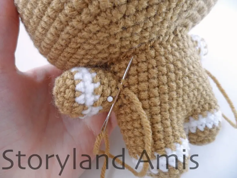 A close-up of a crocheted arm being sewn onto the body of an amigurumi gingerbread boy using a silver tapestry needle.