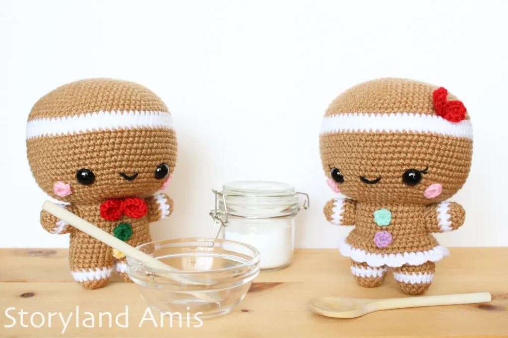 An amigurumi gingerbread man and a gingerbread girl stand on a wooden table surrounded by cooking utensils.