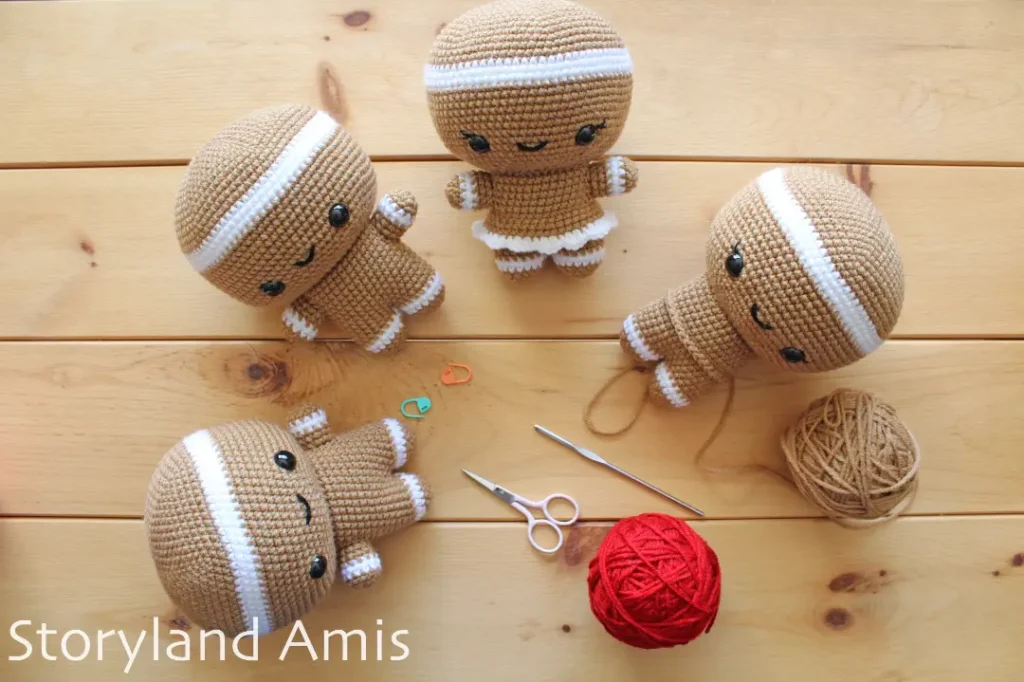 Four partially finished crocheted gingerbread dolls lay on a wooden table in a semicircle. A crochet hook, a pair of scissors, and a red and brown ball of yarn lay scattered on the table.