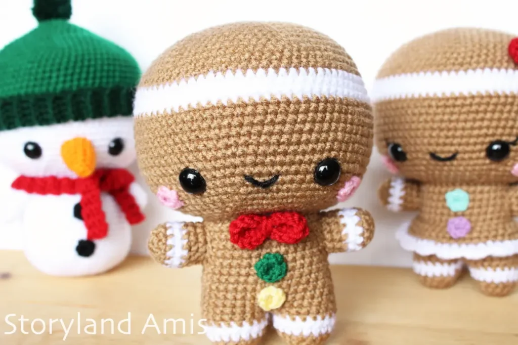 A crocheted gingerbread man stands on a wooden table. Behind him stands a crocheted gingerbread girl and a crocheted snowman.