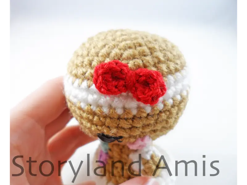 A close-up of the side of the head of an amigurumi gingerbread girl showing a red bow sewn to her head.