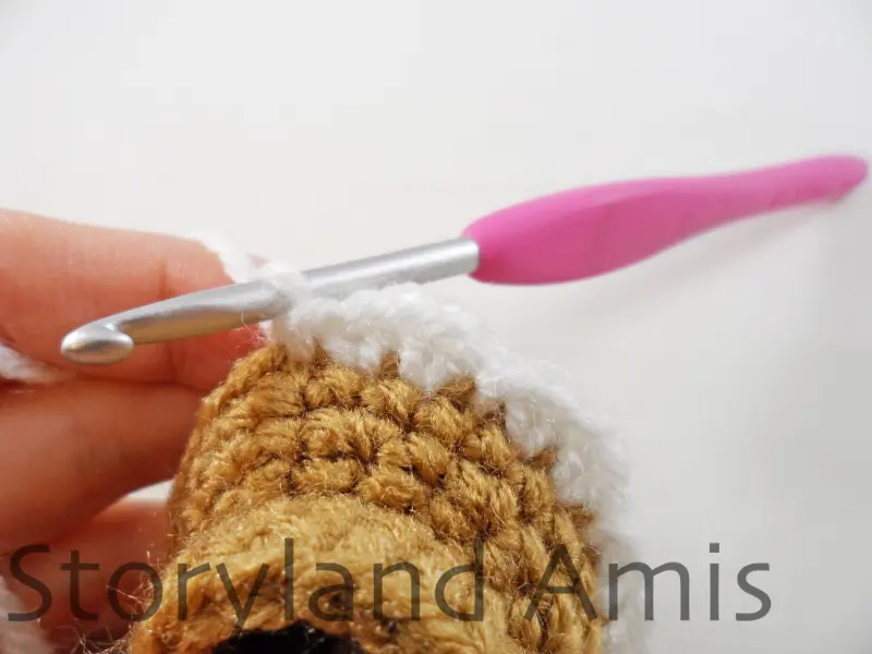 A close-up of the edge of an amigurumi gingerbread girl's skirt. A pink crochet hook is inserted in a stitch on the skirt, showing how to crochet the skirt's border.