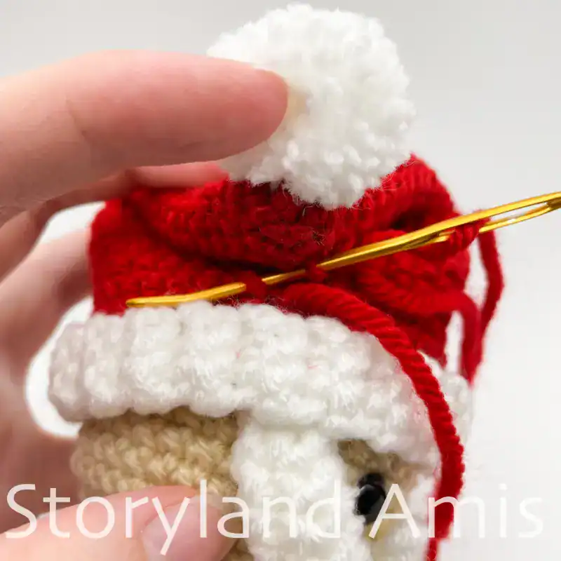 A close-up of a crocheted hat on an amigurumi Santa Claus. A hand uses a tapestry needle to sew the tip of the hat to the side of the hat.