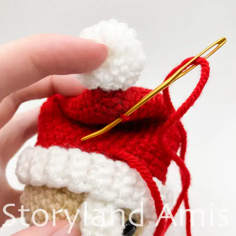 A close-up of a crocheted hat on an amigurumi Santa Claus. A hand uses a tapestry needle to sew the tip of the hat to the side of the hat.