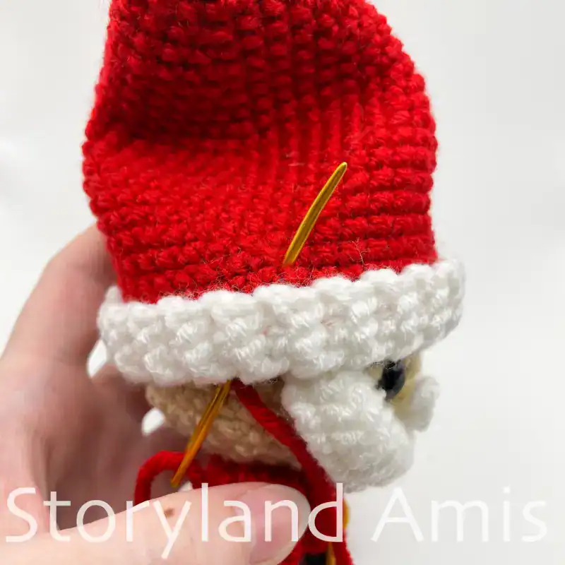 A close-up of the side of a crocheted Santa Claus hat on an amigurumi Santa Claus. A hand holds the doll steady, and a gold tapestry needle is inserted in the base of the hat.
