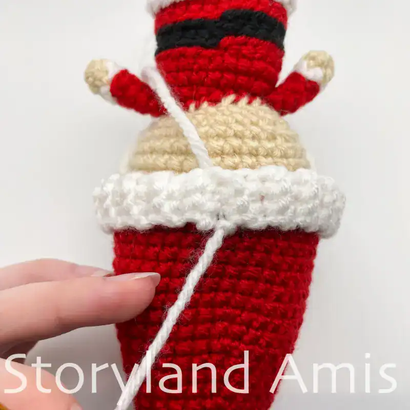The backside of a crocheted Santa Claus doll is shown upside down. Two yarn tails extend from the hat band.
