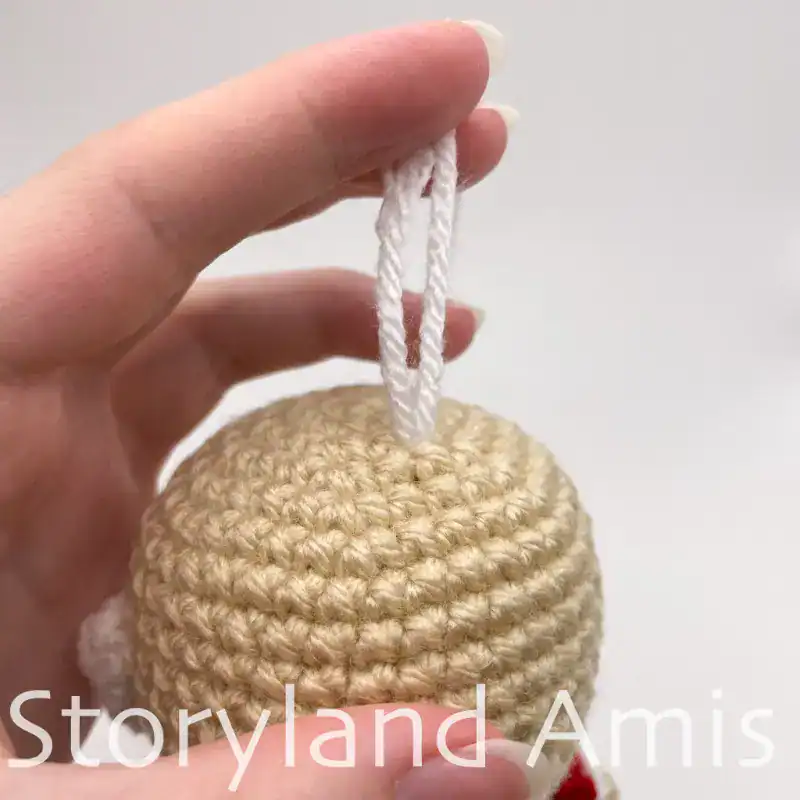 Close-up of a crocheted doll head with two yarn tails coming out of the top of it. A hand holds the two yarn tails straight up.