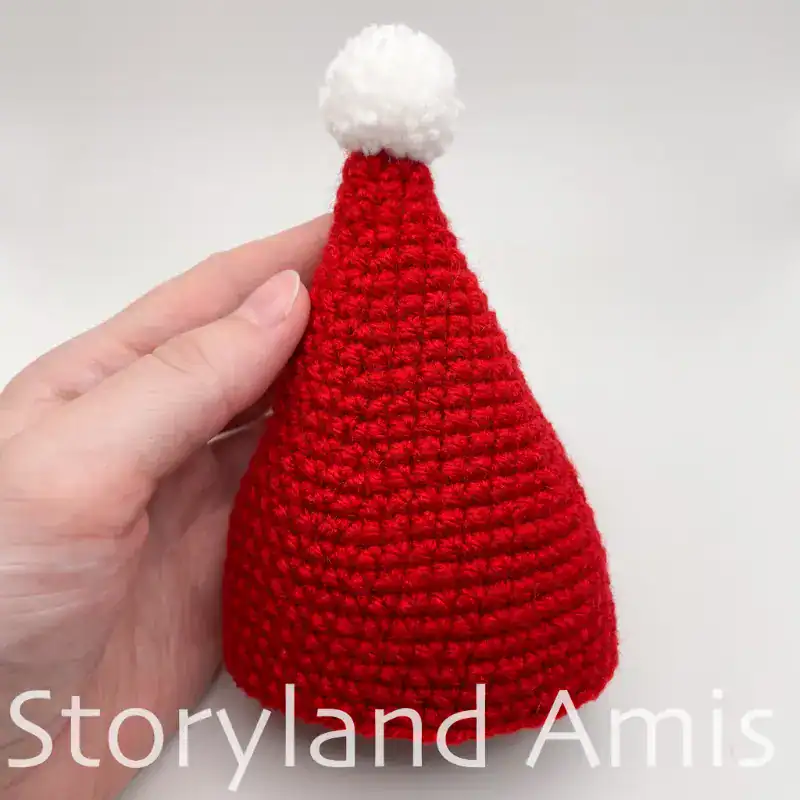 A hand holds a crocheted red hat with a white pom pom on top.