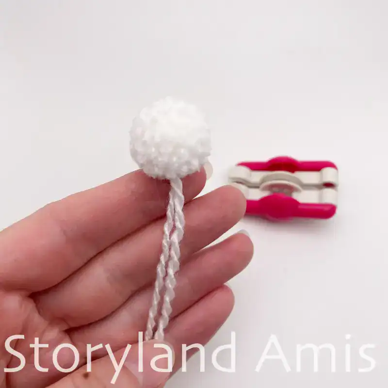 A hand holds a small white pom pom with two yarn tails coming out of it. A pink pom pom maker sits in the background.