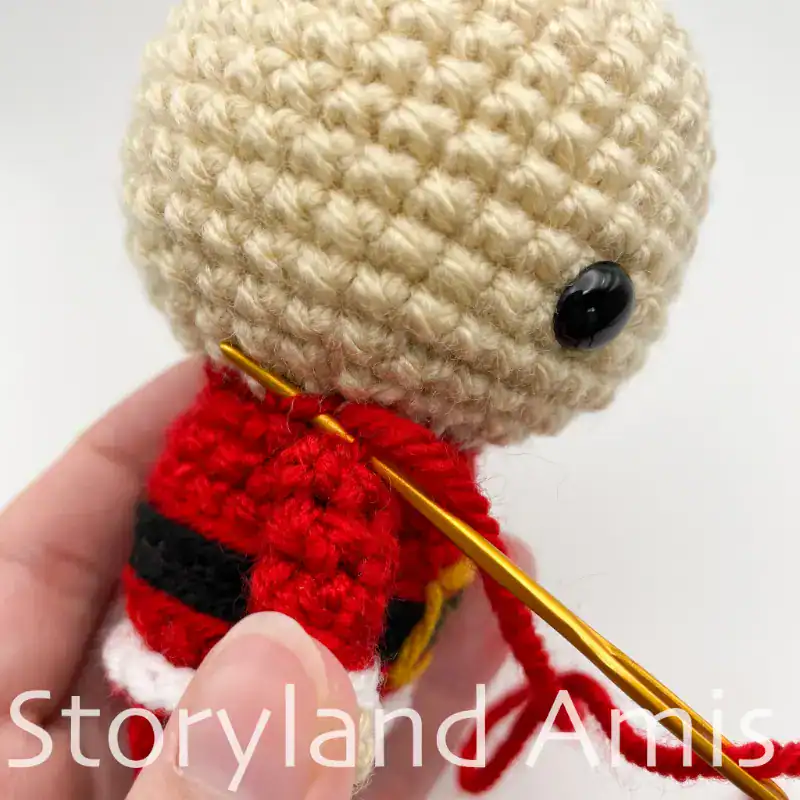 A hand uses a tapestry needle to demonstrate how to sew an arm to the body of an amigurumi Santa Claus doll.