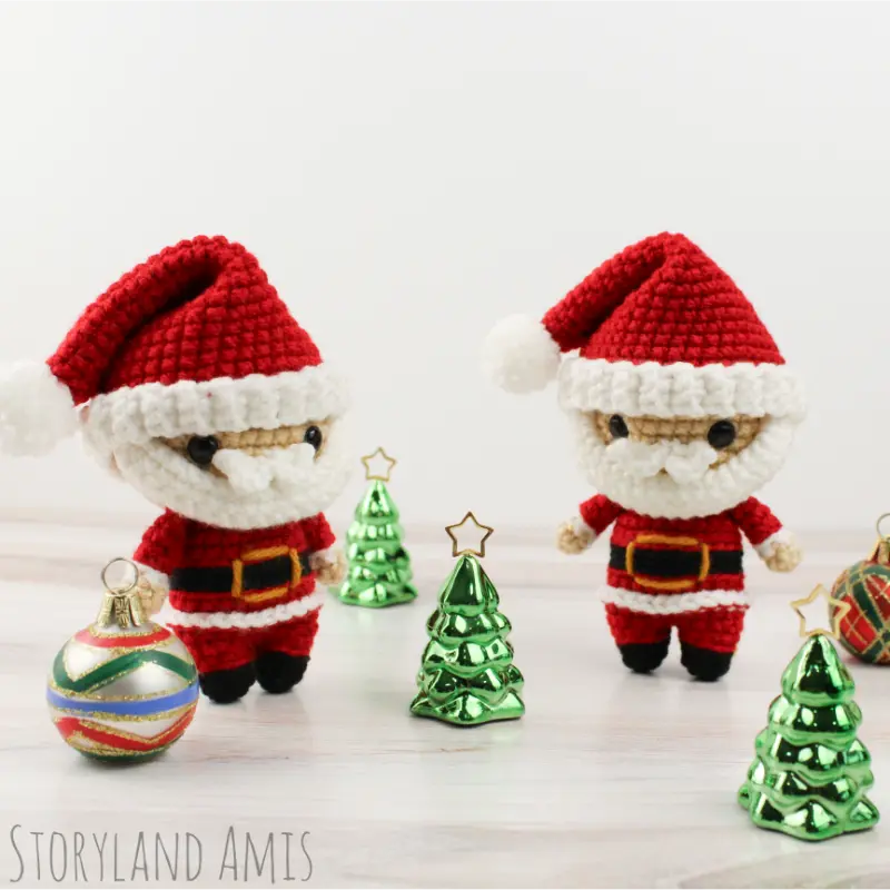 Two mini Santa Claus amigurumi dolls stand on a wooden table. Small metal Christmas trees and ornaments surround them.