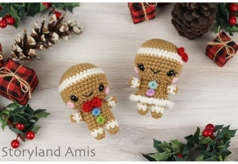 A crocheted gingerbread boy and girl lay on a wooden background surrounded by pinecones, holly leaves, and small plaid wrapped presents.