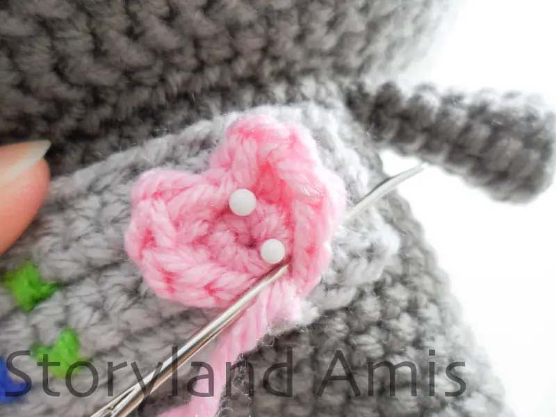 A close-up showing a small pink crocheted heart being sewn onto the front of an amigurumi robot's body.