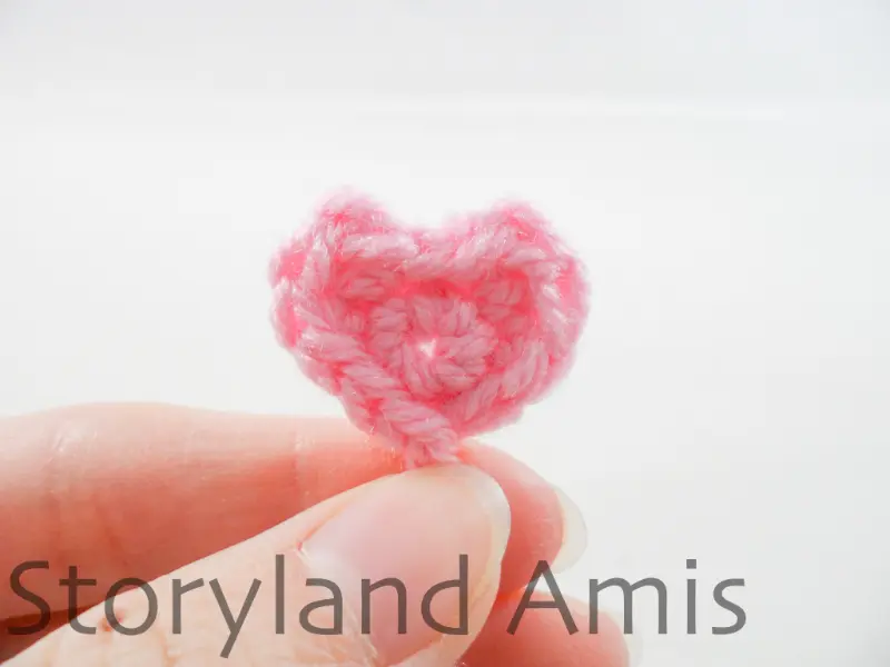 A hand holds up a small pink crocheted heart.
