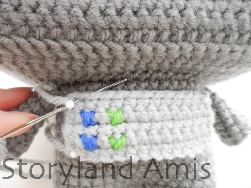 A close-up of an amigurumi robot's chest as a gray rectangular panel is being sewn to it using a metal tapestry needle.