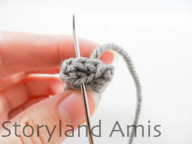 A hand demonstrates how to sew the open ends closed on a crocheted robot arm using a metal tapestry needle.