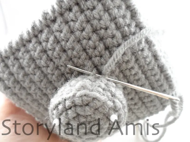 A close-up showing a round gray crocheted piece being sewn to the side of an amigurumi robot's head.