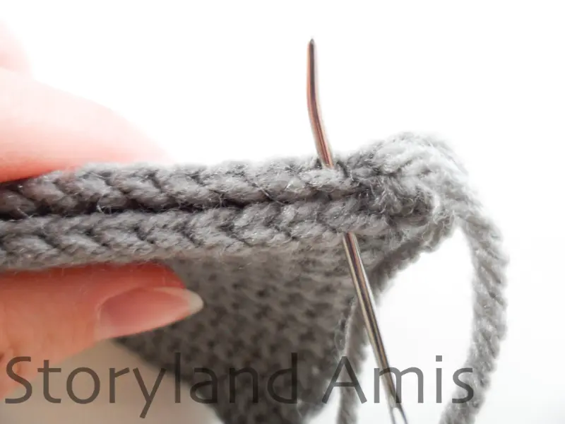 A close-up of two gray crocheted rectangular panels being sewn together with a tapestry needle. A hand holds the two panels together.